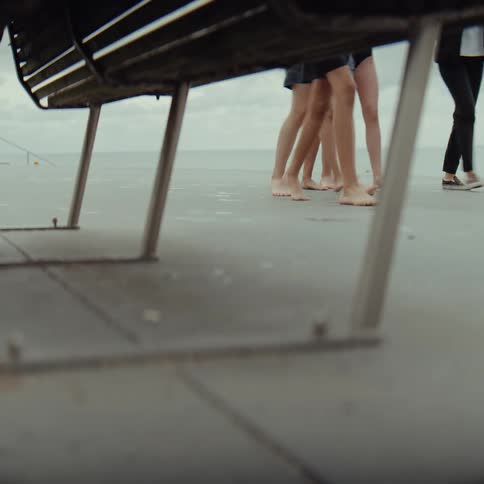 People walking barefoot and in shoes along a seaside promenade viewed from beneath a bench