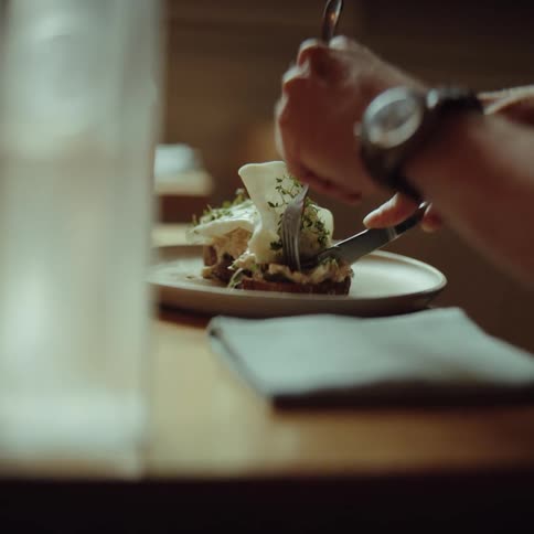 A person uses a silver fork and knife to cut into a poached egg topped with green herbs on a piece of toast served on a white plate