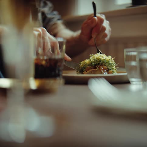 Person using a fork to eat a plated dish at a table with glasses and a drink