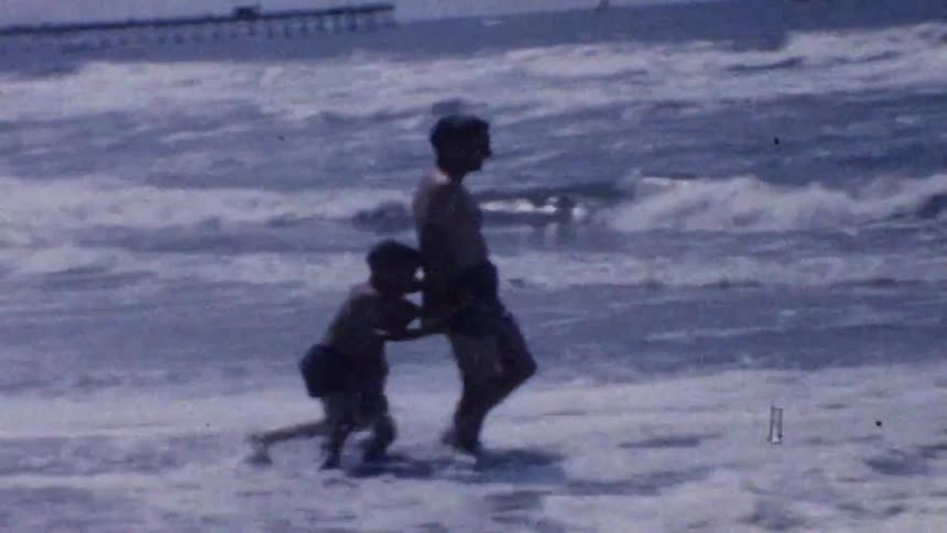 The video shows a vintage grainy home movie of a man and a young boy playing in the ocean wavesthe child clings to the mans waist as they wade through the churning white surf with a pier visible in the distant backgroundwould you like me to descri
