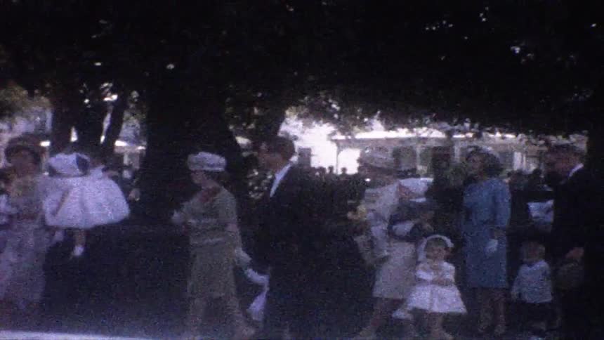 A crowd of people dressed in formal midcentury attire walks through a shaded outdoor areaseveral young children in white dresses are visible among the adults near a large tree