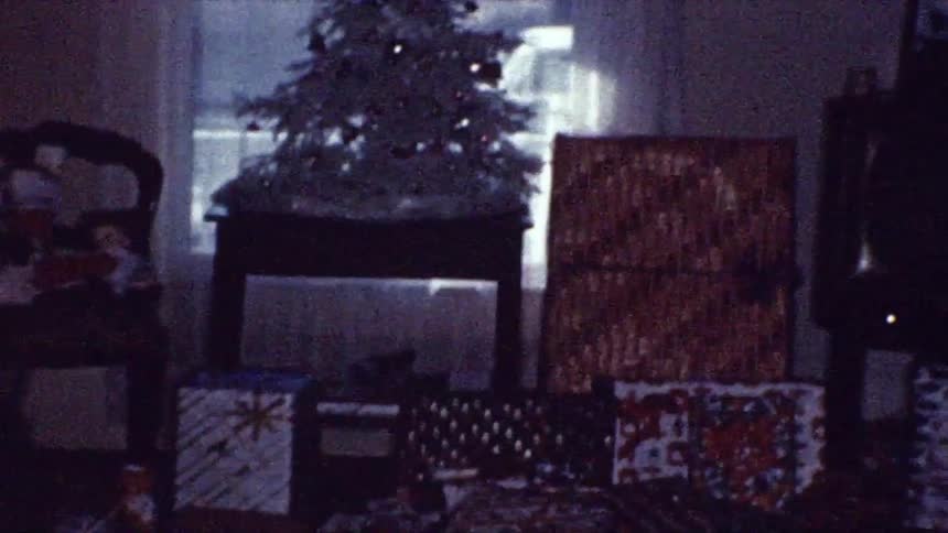A decorated christmas tree stands on a table surrounded by wrapped gifts in a dimly lit room