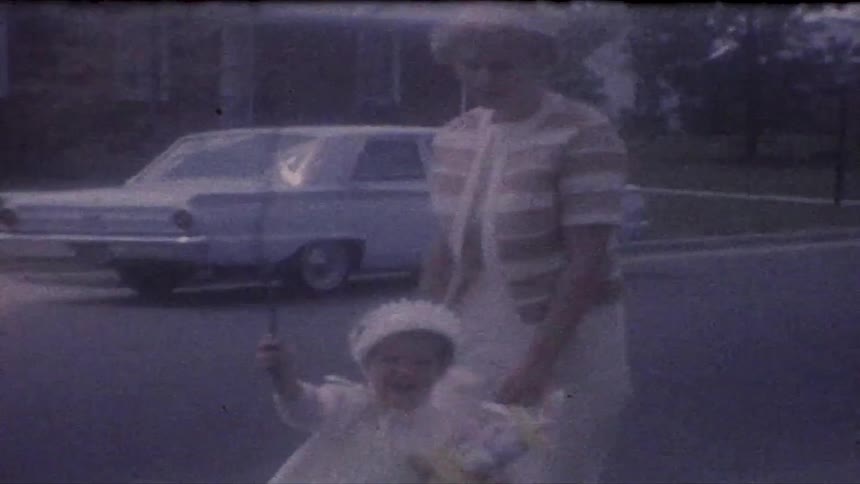 A woman in a striped cardigan supervises a toddler in a white bonnet as the child walks along a suburban curb holding a stuffed bunny and a thin stick with a vintage blue car parked on the street in the background