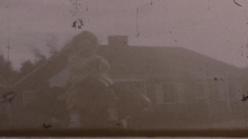 A man in a dark jacket squats while carrying a small child in a yellow shirt on his shoulders in a backyard with a suburban ranchstyle house and trees visible in the background