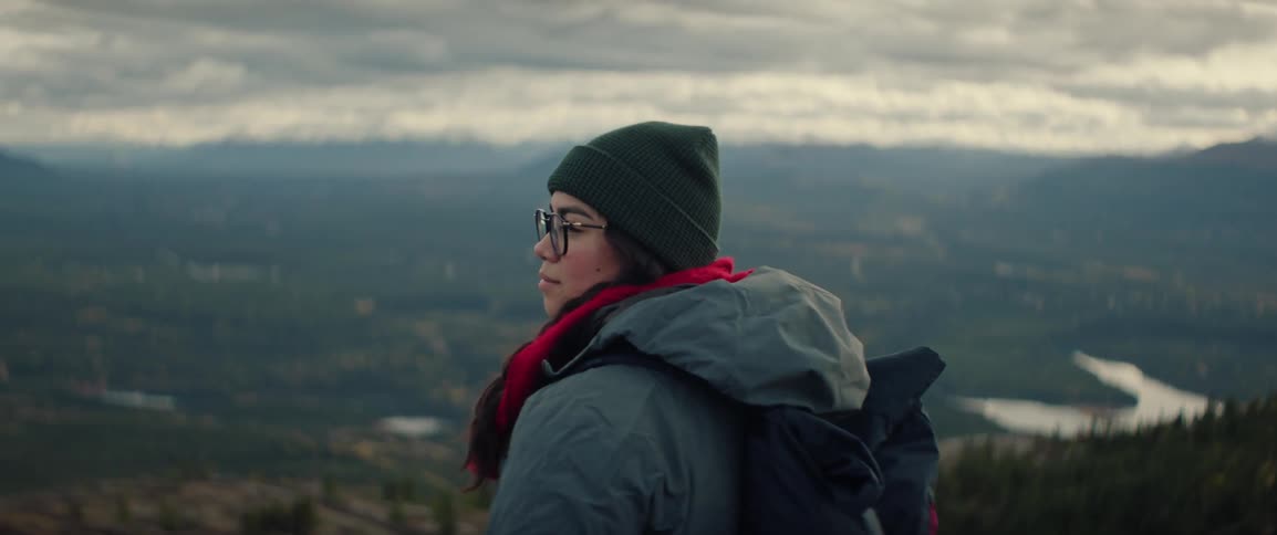 A young woman standing on a mountaintop overlooks a vast valley with winding rivers and distant peaks