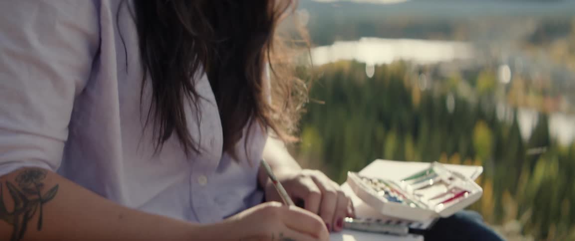 A woman creates a watercolor painting in her sketchbook while sitting outdoors in daylight