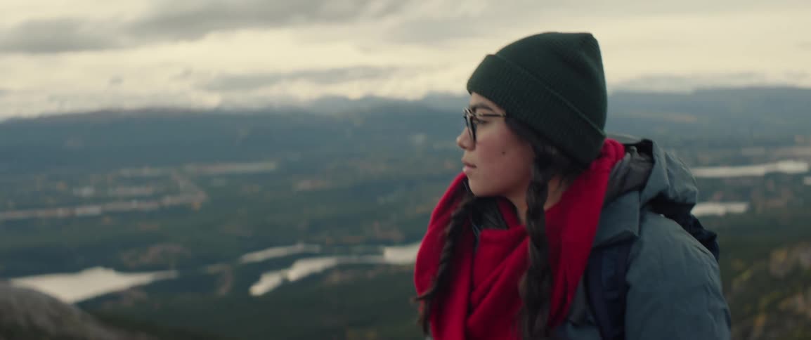 A woman with dark braids wearing a green beanie walks up a mountain overlooking a vast landscape and distant mountains