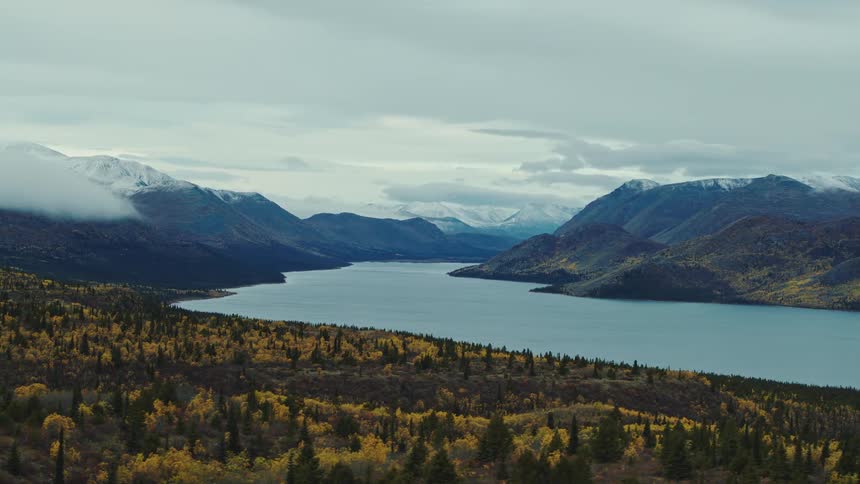 Vast lake between mountain ranges with autumn toned forest under a cloudy sky