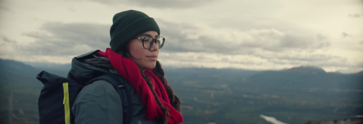 Woman wearing a beanie glasses a backpack and a scarf at a viewpoint with a mountainous landscape in the background