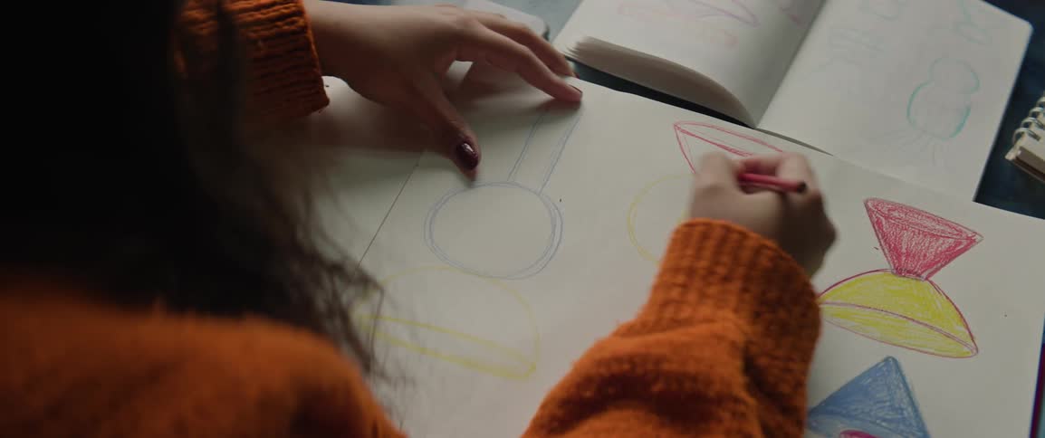 A girl wearing an orange knit sweater uses a red colored pencil to draw geometric shapes on a large white sketchpad