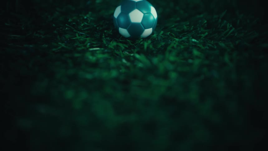 A blue and white soccer ball sits on a green artificial turf field under dark moody lighting