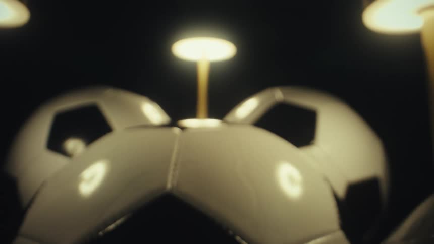A closeup moving shot captures several black and white soccer balls resting under warm circular desk lamps against a dark background highlighting the glossy texture and stitching of the equipment