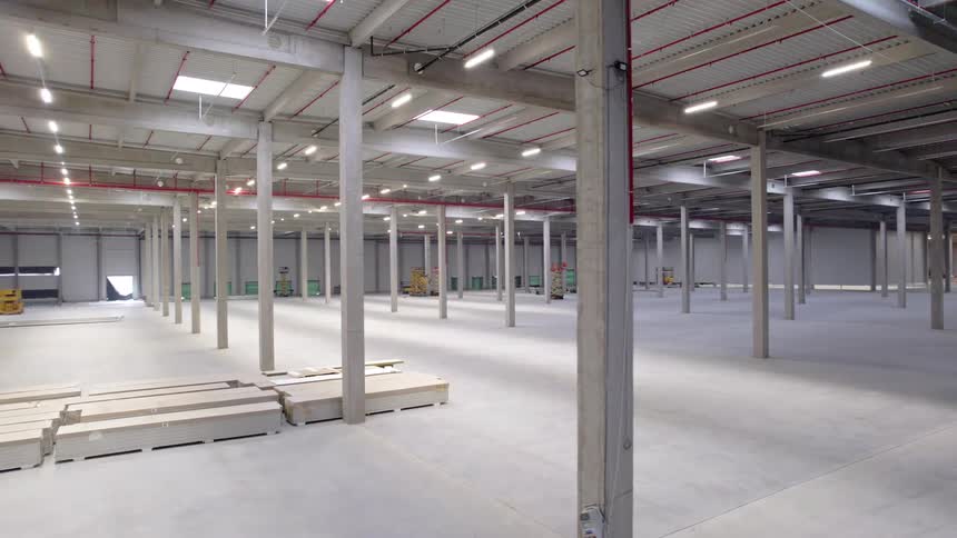 A wide shot shows the interior of a massive empty warehouse with concrete floors and numerous support pillars under bright industrial lighting
