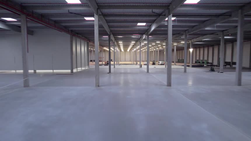 A massive empty warehouse under construction showcasing the expansive concrete floor rows of support pillars and high ceilings while workers and service vehicles occupy small sections of the far background area