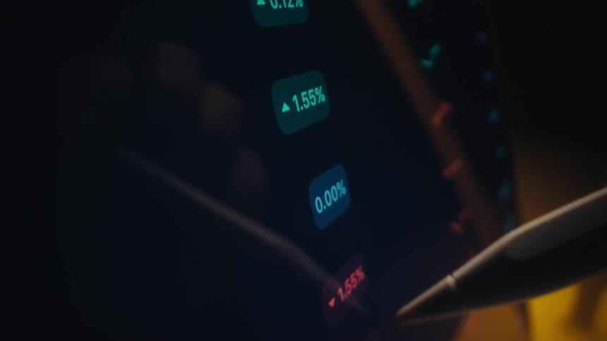 A person uses a white stylus to scroll through a list of stock market percentages and financial data on a dark tablet screen highlighting various red and green price fluctuations in a low light setting