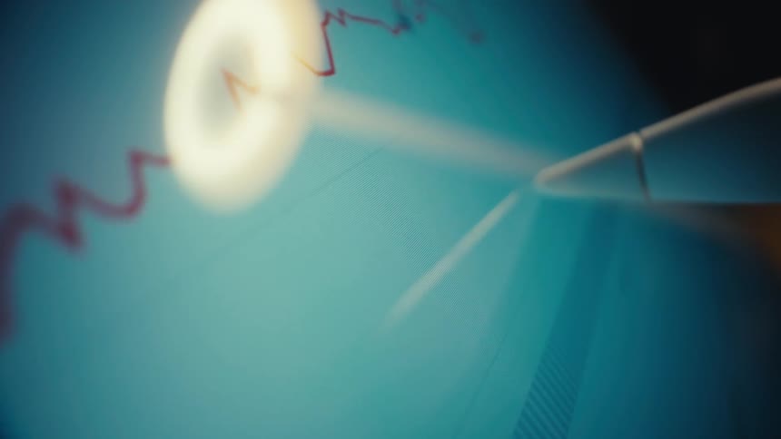A closeup shot of a digital screen displays a fluctuating red line graph representing market data while a white stylus pen moves across the surface near a circular light reflection