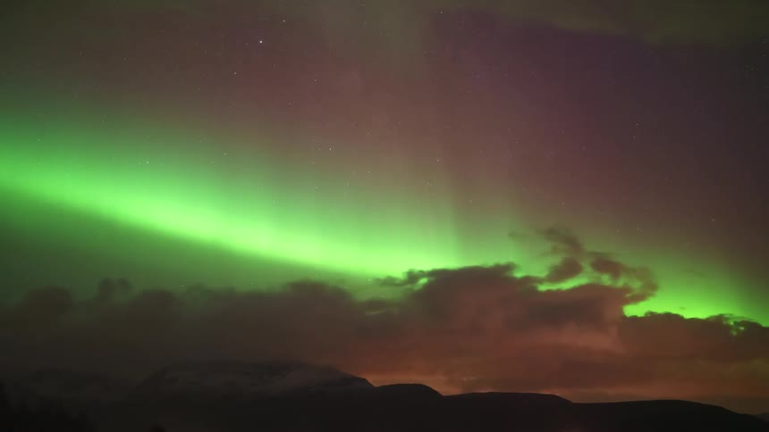 The glowing green lights of the aurora borealis move above clouds over the landscape