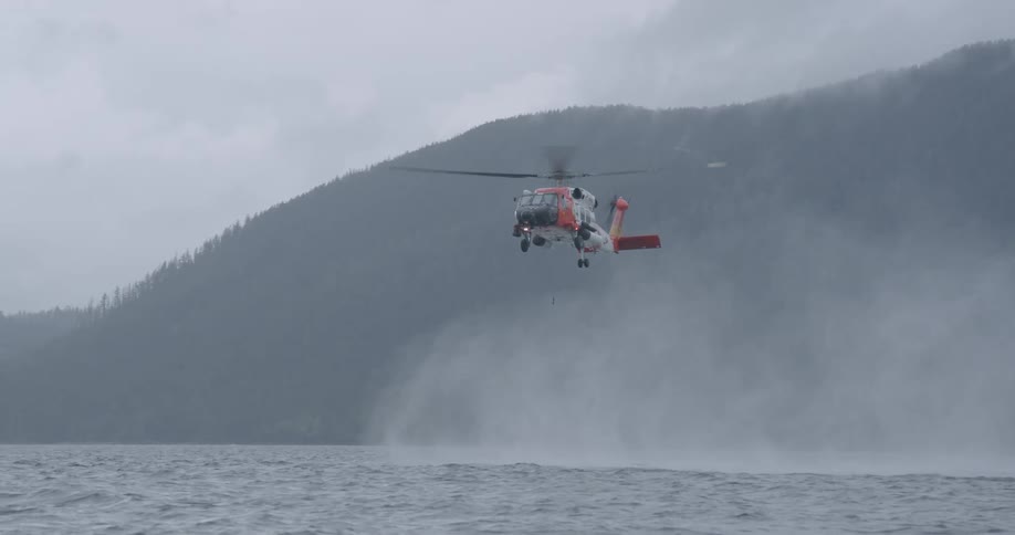 A coast guard helicopter hovers low over a choppy body of water creating a large spray of mist with its rotors while a dark forested mountain rises in the background under a grey overcast sky