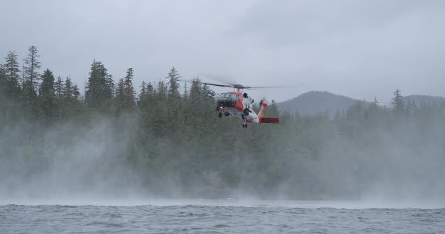 A red and white rescue helicopter hovers low over choppy water creating a strong downdraft mist against a backdrop of a dense pine forest under an overcast sky