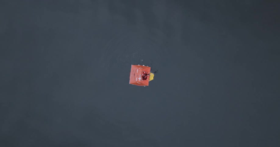 A top down shot shows a man rowing on an orange square life raft floating on a dark vast body of water