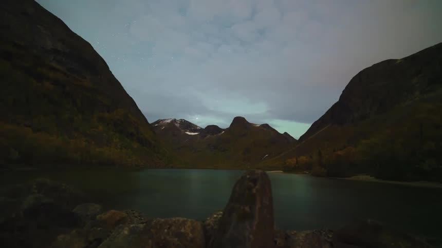 A timelapse video shows a dark valley at night where clouds drift over a calm lake nestled between steep mountains while stars move across the sky above