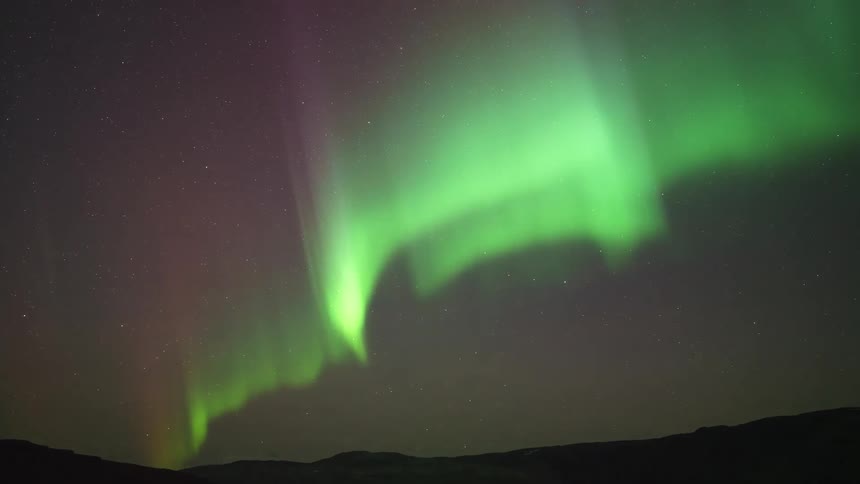 Vibrant green and purple aurora borealis shimmer and dance across a dark starfilled night sky above the silhouettes of distant lowlying mountains and a vast arctic horizon