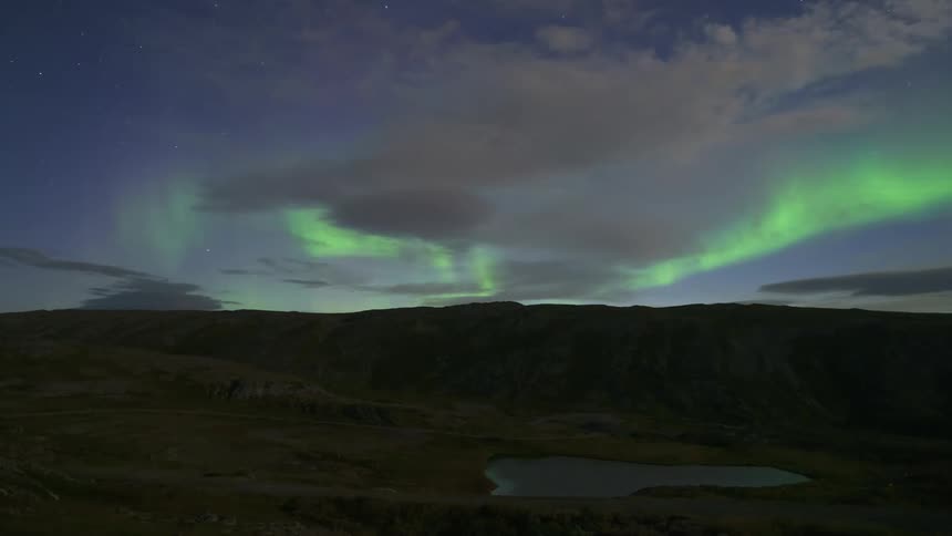 A timelapse captures bright green northern lights shimmering and shifting across a dark star filled sky above a vast mountain landscape with a small lake in the foreground