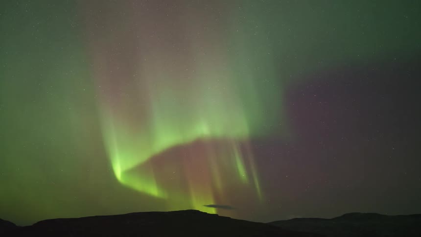 The aurora borealis dances across a starry night sky in shades of green and purple over the dark silhouette of a mountain range