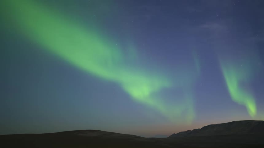 A timelapse video shows the green aurora borealis dancing across a dark blue night sky over a desolate landscape of rolling hills and distant mountains as the stars shift slowly in the background