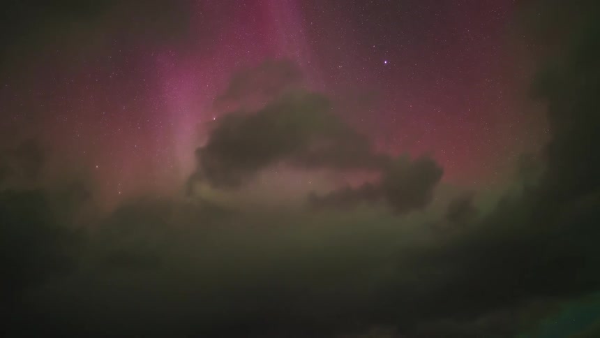 A stunning timelapse reveals vibrant green and pink aurora borealis dancing in the starry night sky as dark clouds drift swiftly across the celestial display