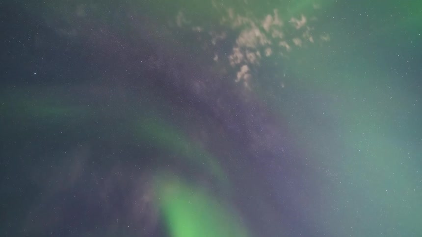 A timelapse captures vibrant green and purple northern lights swirling and dancing across a star filled night sky while the camera points directly upward toward the zenith