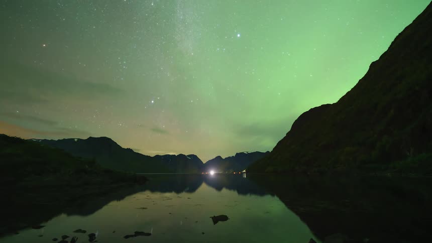 A timelapse captures vibrant green northern lights shimmering and shifting across a star filled night sky while reflecting on the surface of a calm lake nestled between dark silhouettes of large mountains