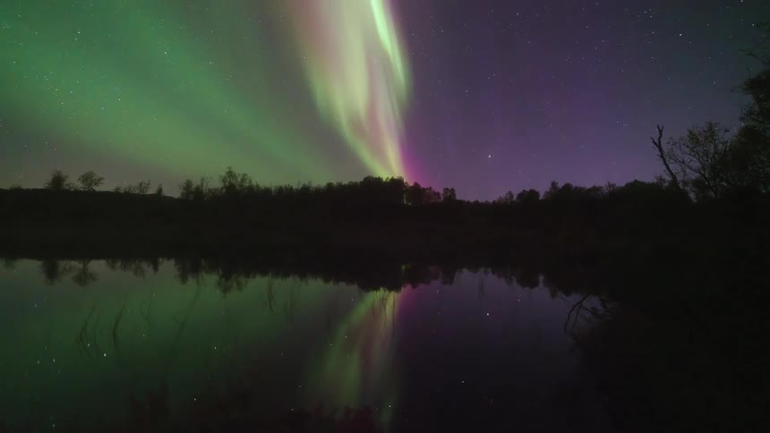 A lake sits quietly at night surrounded by dark tree lines while a green light hovers in the sky above and reflects faintly on the water