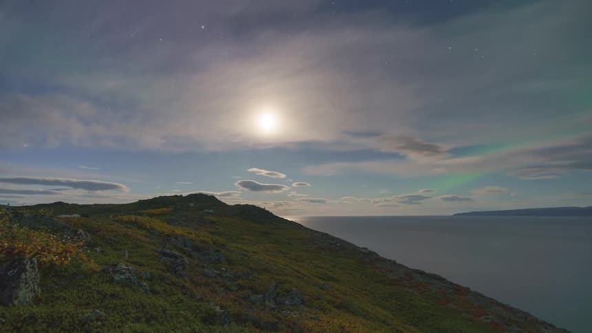 A stunning timelapse transitions from a colorful sunset to a starry night where the moon rises to cast its light on the sea and reveal the faint dance of the aurora