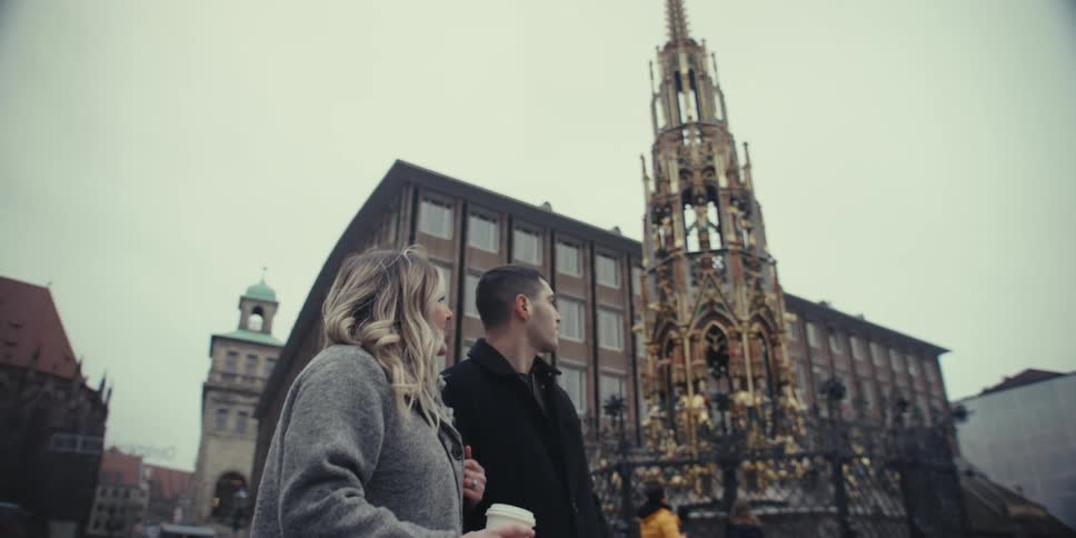 A couple walks together through a historic european square admiring the schner brunnen fountain and the frauenkirche in nuremberg they talk and smile while holding coffee cups enjoying a gray overcast day in the city center