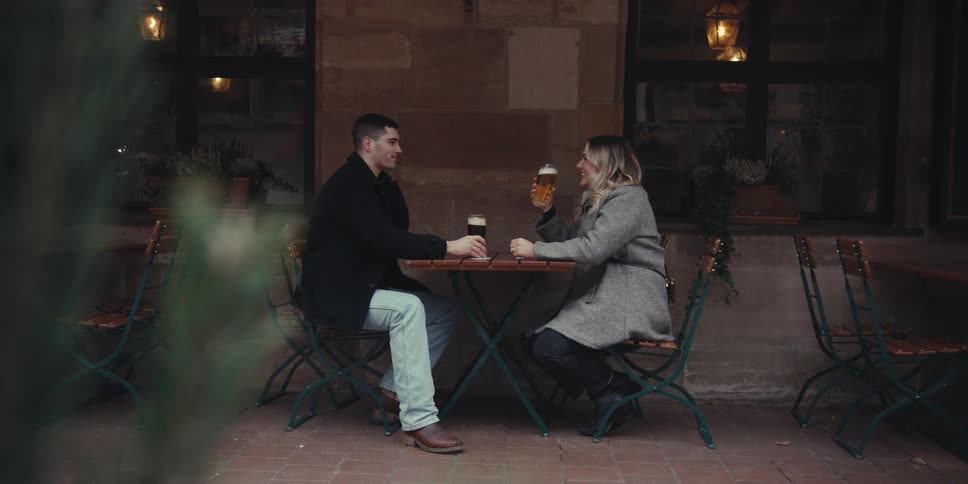 A couple sits at an outdoor caf table smiling at each other while enjoying drinks in a cozy warmly lit setting