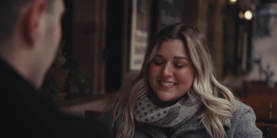 Man and woman sitting at an outside table as they smile toasting with their beers then they kiss