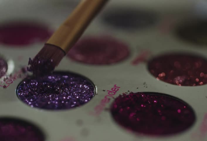 A makeup brush gently presses into a pan of sparkling purple glitter within a cosmetics palettethe tool lifts a small cluster of the shimmering particles revealing the reflective texture of the surrounding pink and magenta shades