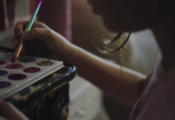 A person uses a multi colored brush to dip into a glittery makeup palette of various colors