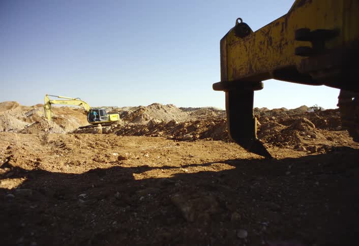 Heavy construction equipment sits across a dusty excavation site under a clear blue sky with a large metal blade in the foreground and an excavator working in the distance