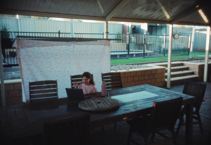 A young child sits at a large outdoor table under a covered patio focused on a laptop waving her hand in a quiet backyard setting