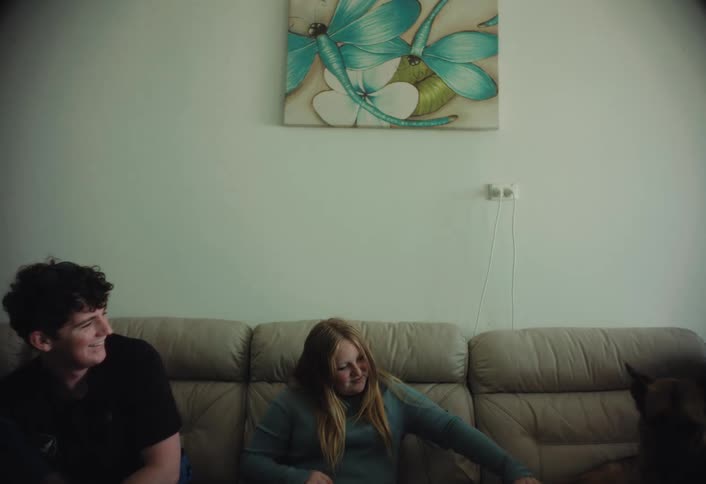 Two young people sit on a couch and smile while interacting with a large dog the scene is set in a living room featuring a lightcolored sofa and a dragonfly painting on the wall