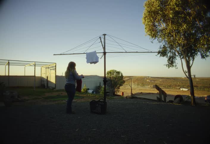 Woman hangs laundry on an outdoor frame while a car drives past in the distance on a sunny day