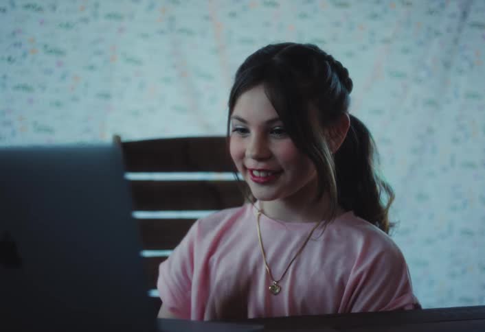 A young girl sits at a table in front of a laptop waving and smiling