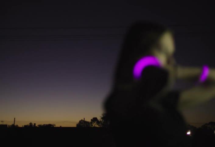A blurry woman wearing glowing purple bracelets dances at night against a dark purple and orange twilight sky