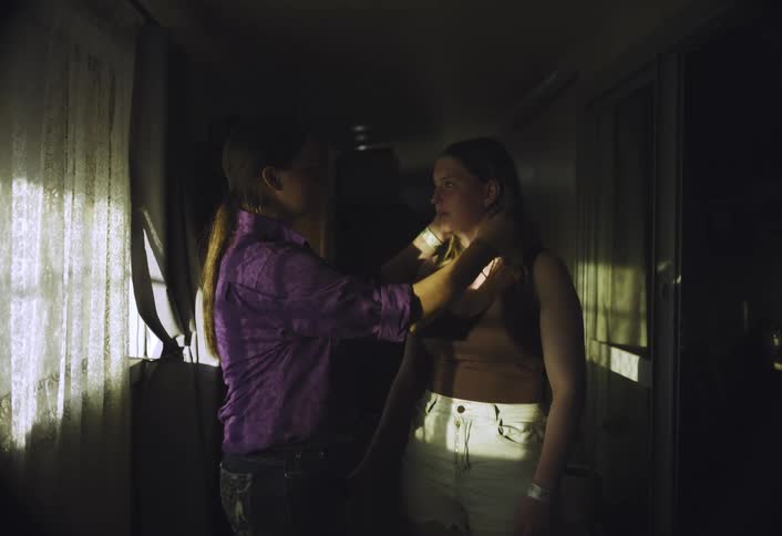 Two women talk in a dark room illuminated by window light mostly falling on the woman wearing white shorts