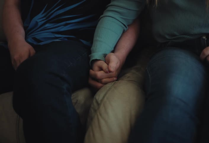 A couple sits close together on a couch their hands intertwined in a gentle hold the low lighting emphasizes the quiet intimate connection shared between the two individuals