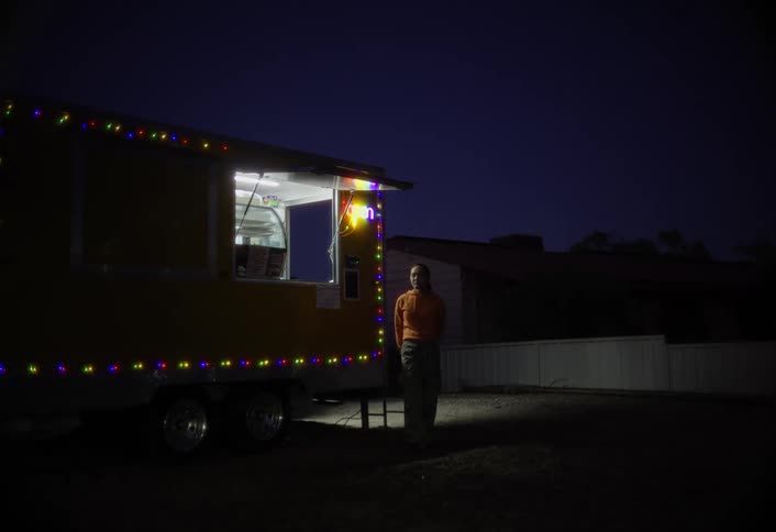 A food truck vendor stands outside waiting for customers under colorful lights at night
