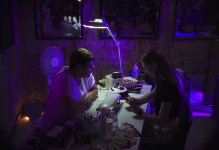 Two people perform a manicure procedure at a table lit with purple light