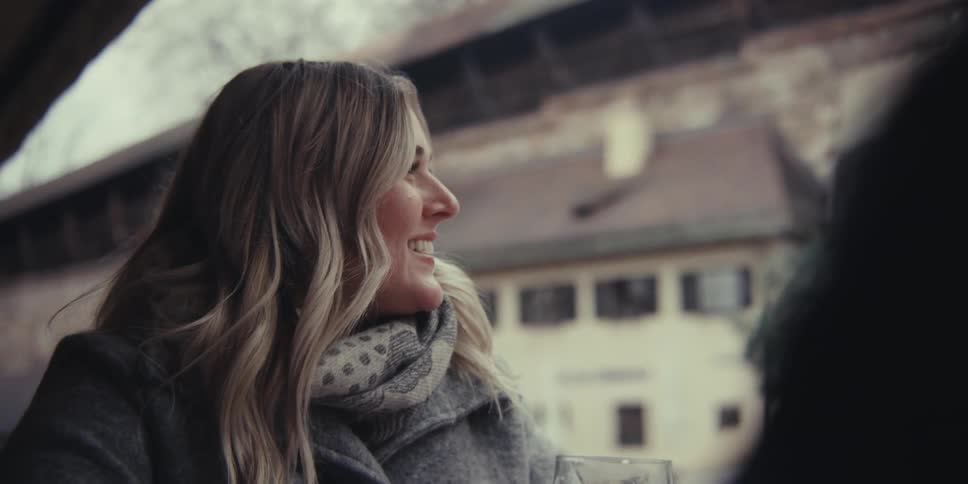 A blonde woman wearing a winter coat and scarf laughs and talks with a companion at an outdoor cafe before taking a sip of her beer against a backdrop of historic buildings
